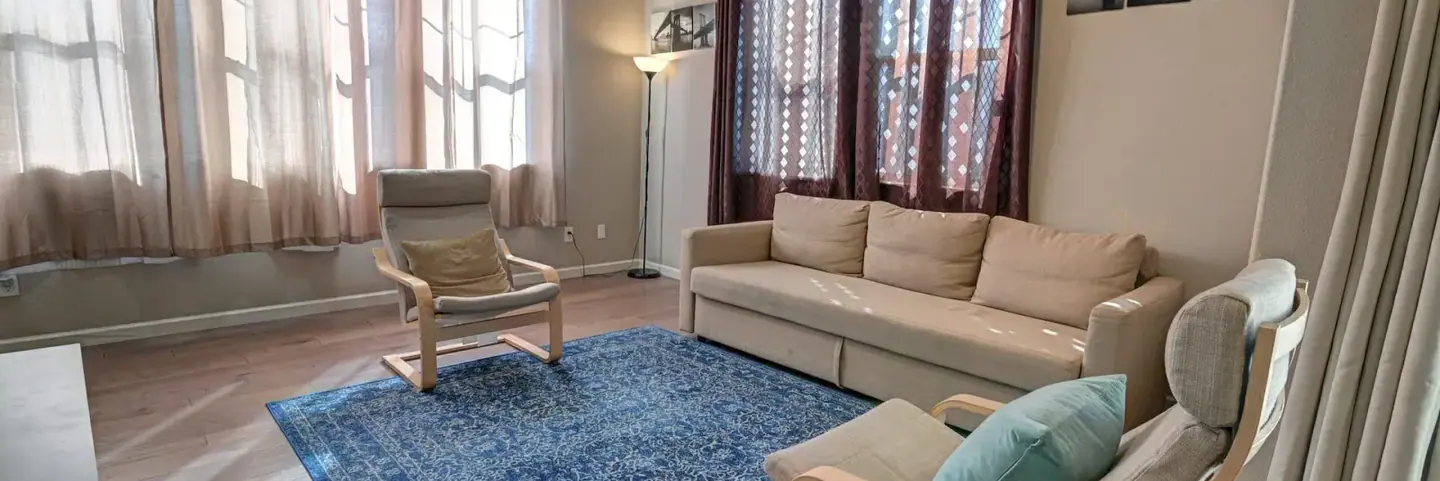 Living room with a beige sofa, wood-frame accent chairs, and framed black-and-white New York City photos on the wall.