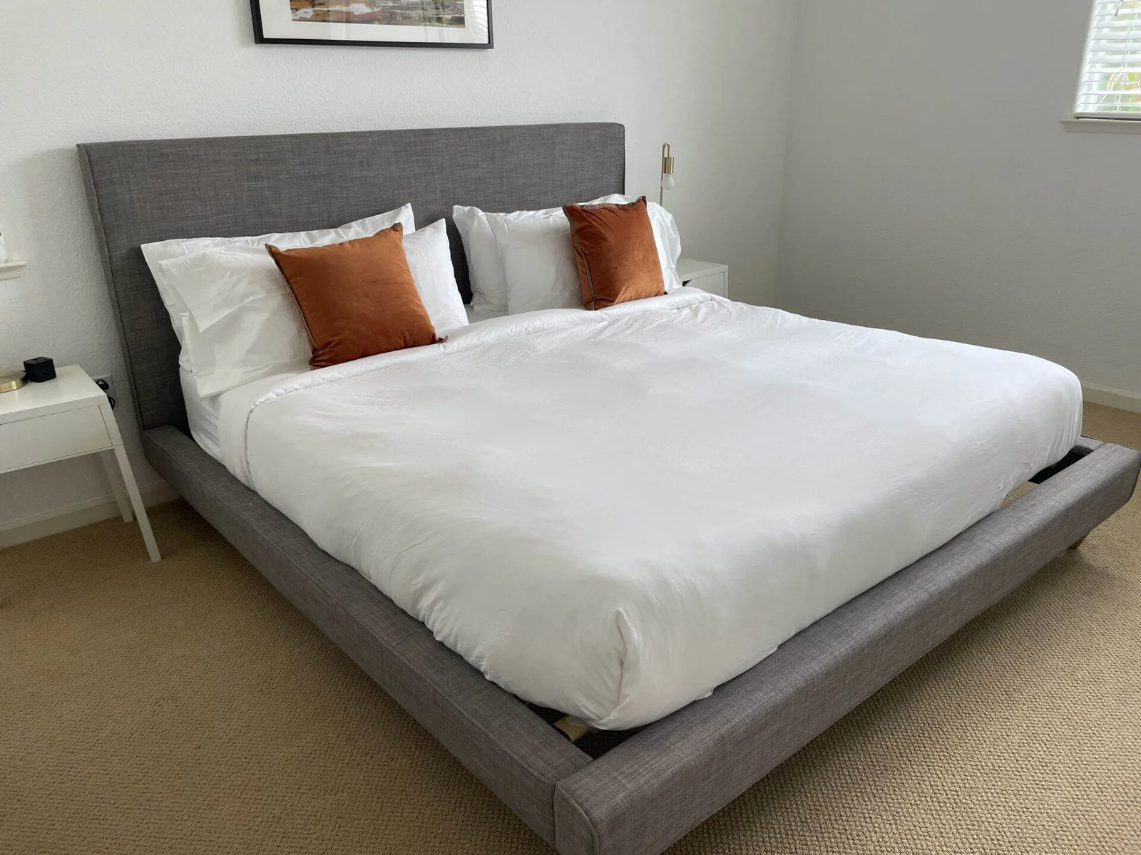 Made bed with white sheets, white pillows, and two rust-orange accent pillows against a gray upholstered headboard.