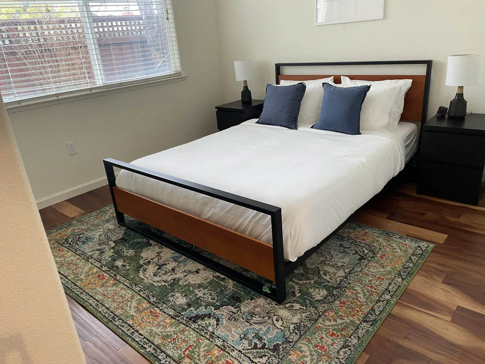 Bedroom with a wood-and-black-metal bed frame, blue throw pillows, and a colorful patterned area rug on wood flooring.
