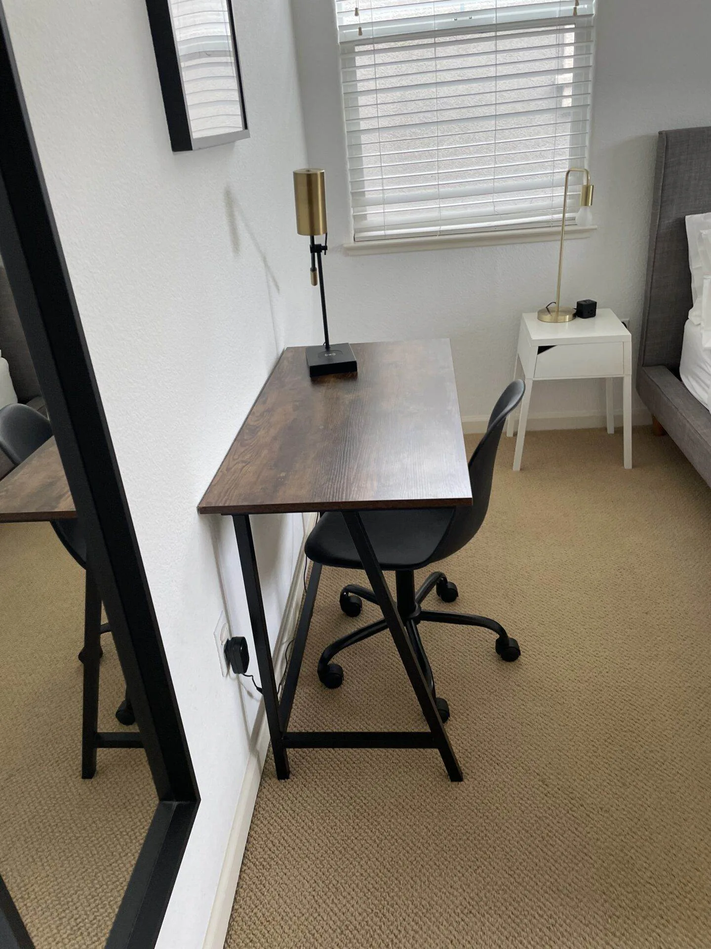 Small wood desk with a brass lamp and black rolling office chair beside a window in a carpeted bedroom.