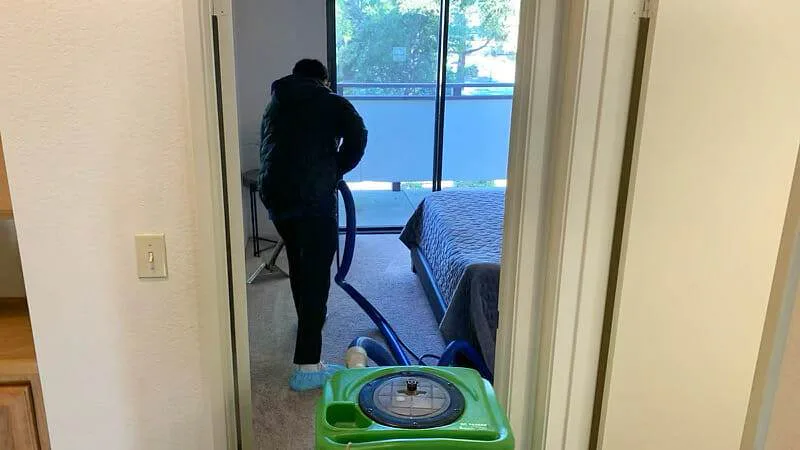 Cleaning technician vacuuming bedroom carpet with a portable carpet extractor near a balcony window in Santa Clara.