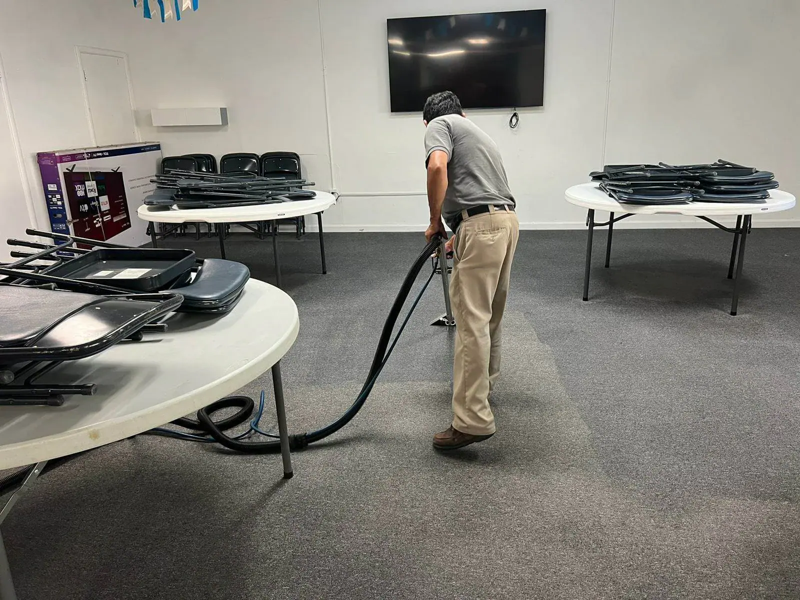 Technician cleaning commercial carpet in an office with tables and stacked folding chairs.
