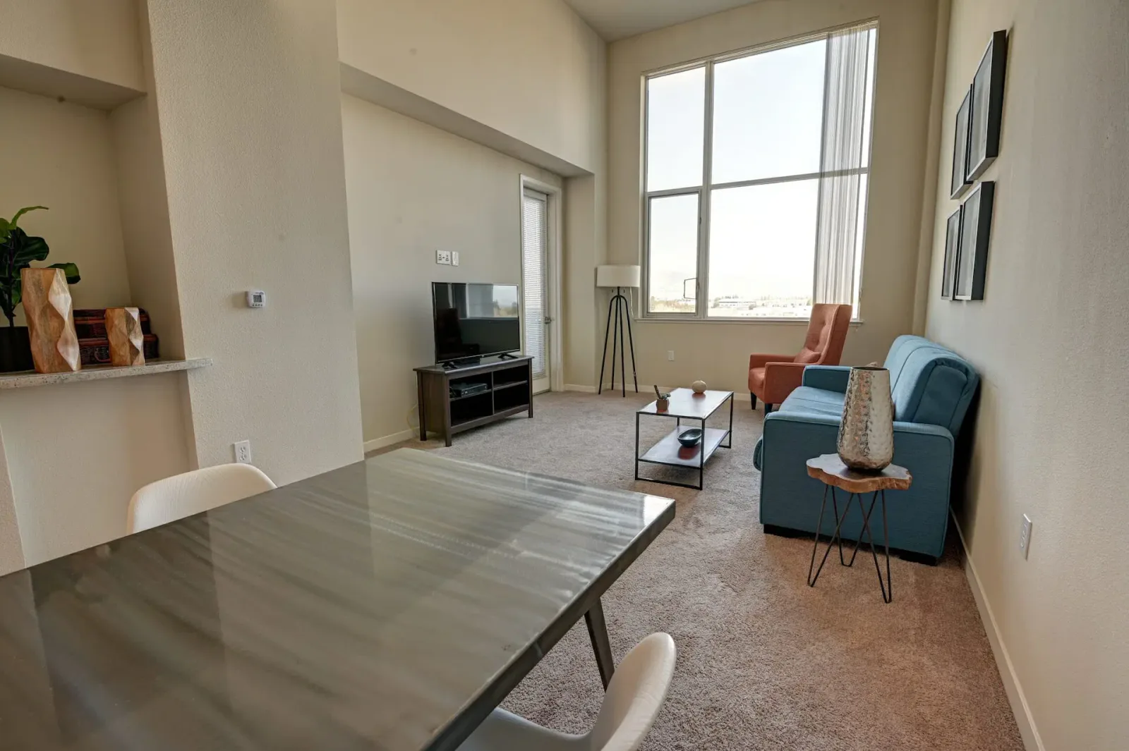 Wide interior view with a glass-top dining table and white chairs in the foreground, leading back to a living room and small TV console.