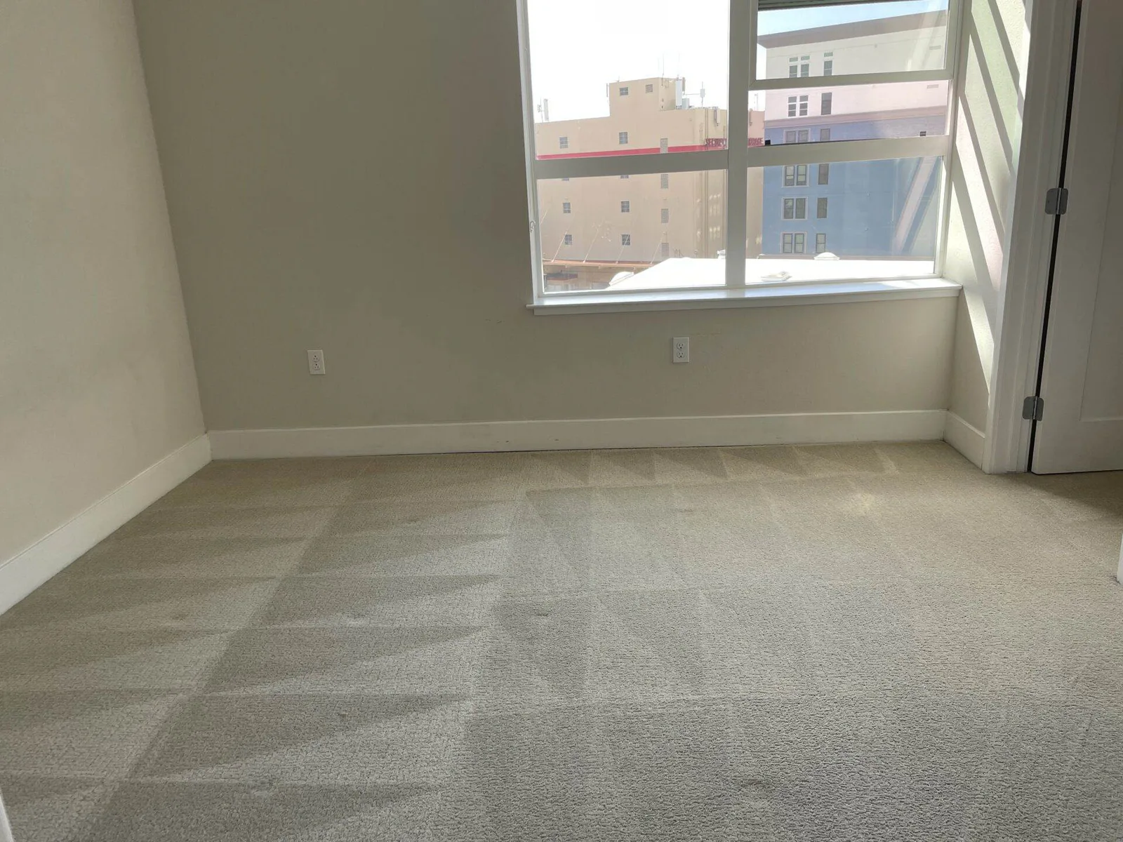 Empty carpeted bedroom with a large window showing a city view, freshly cleaned and vacant.