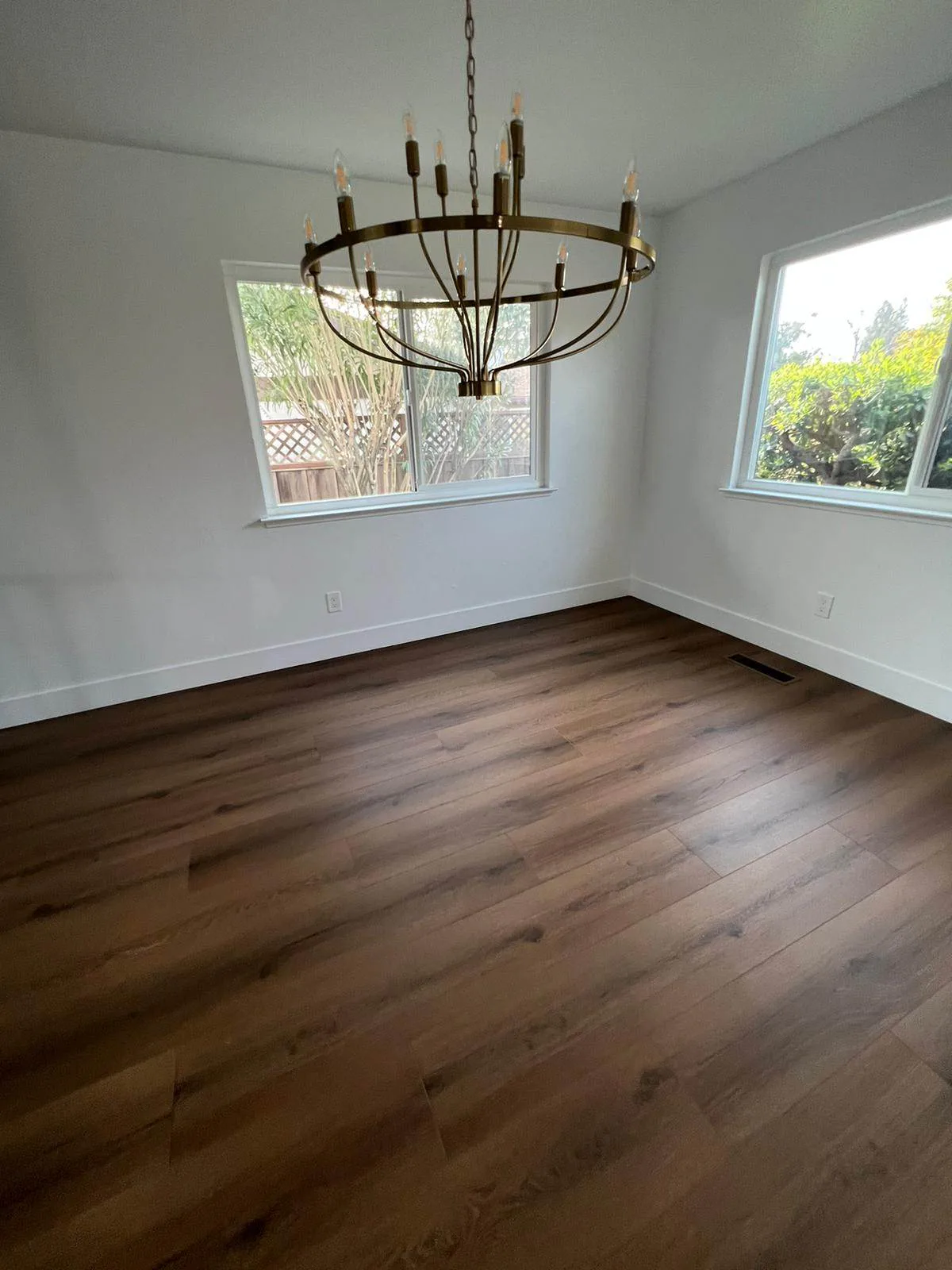 Empty dining room with dark wood flooring, white walls, two windows, and a brass candle-style chandelier.