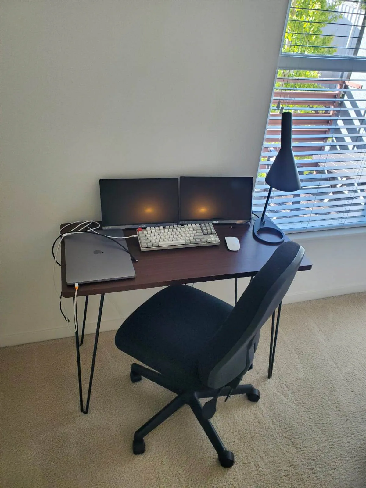 Home office corner with a dark wood desk, dual monitors, mechanical keyboard, and a desk lamp by a window.