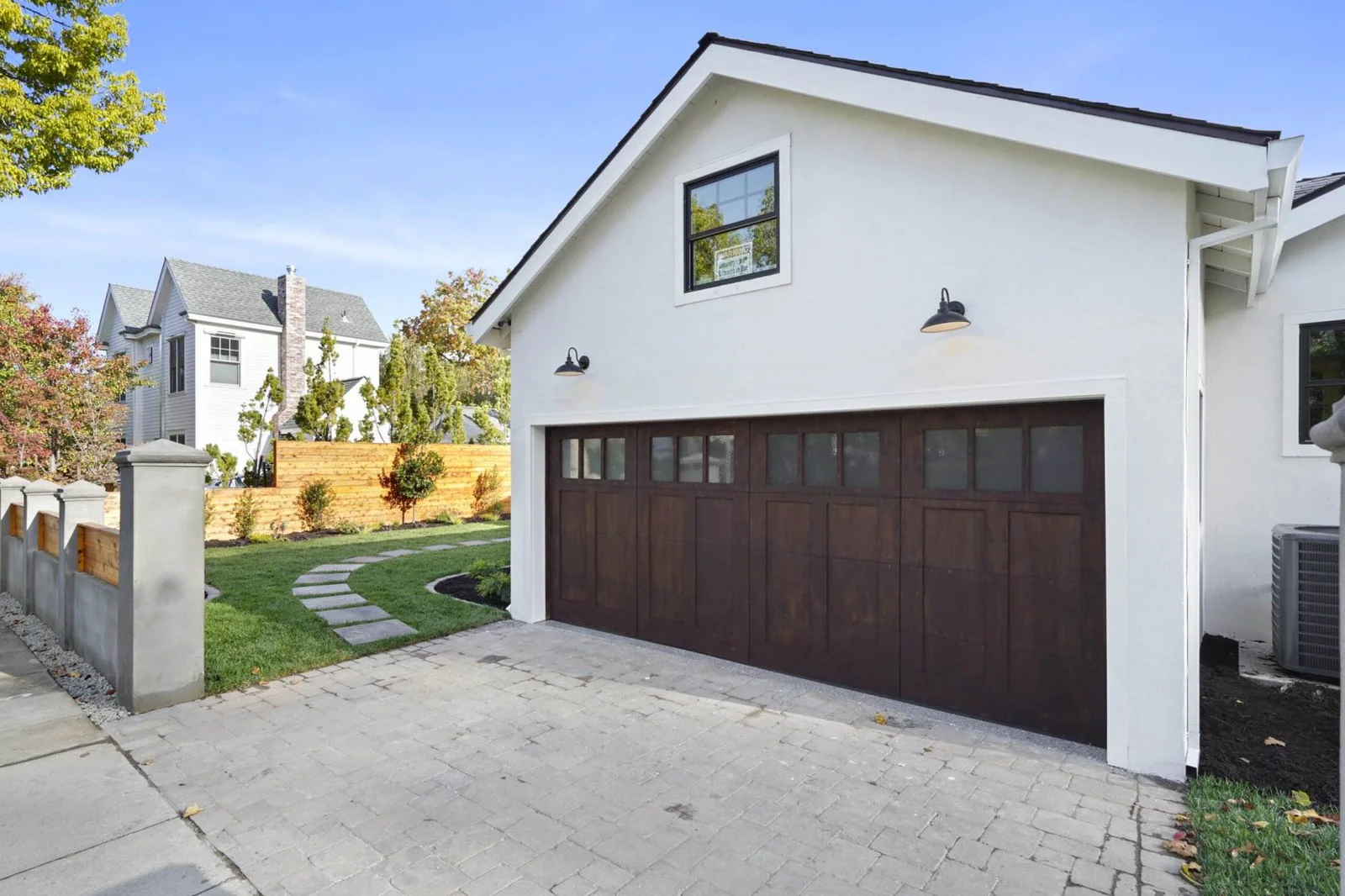 Two-story white house exterior with a dark wood-tone garage door, paver driveway, and landscaped side yard.