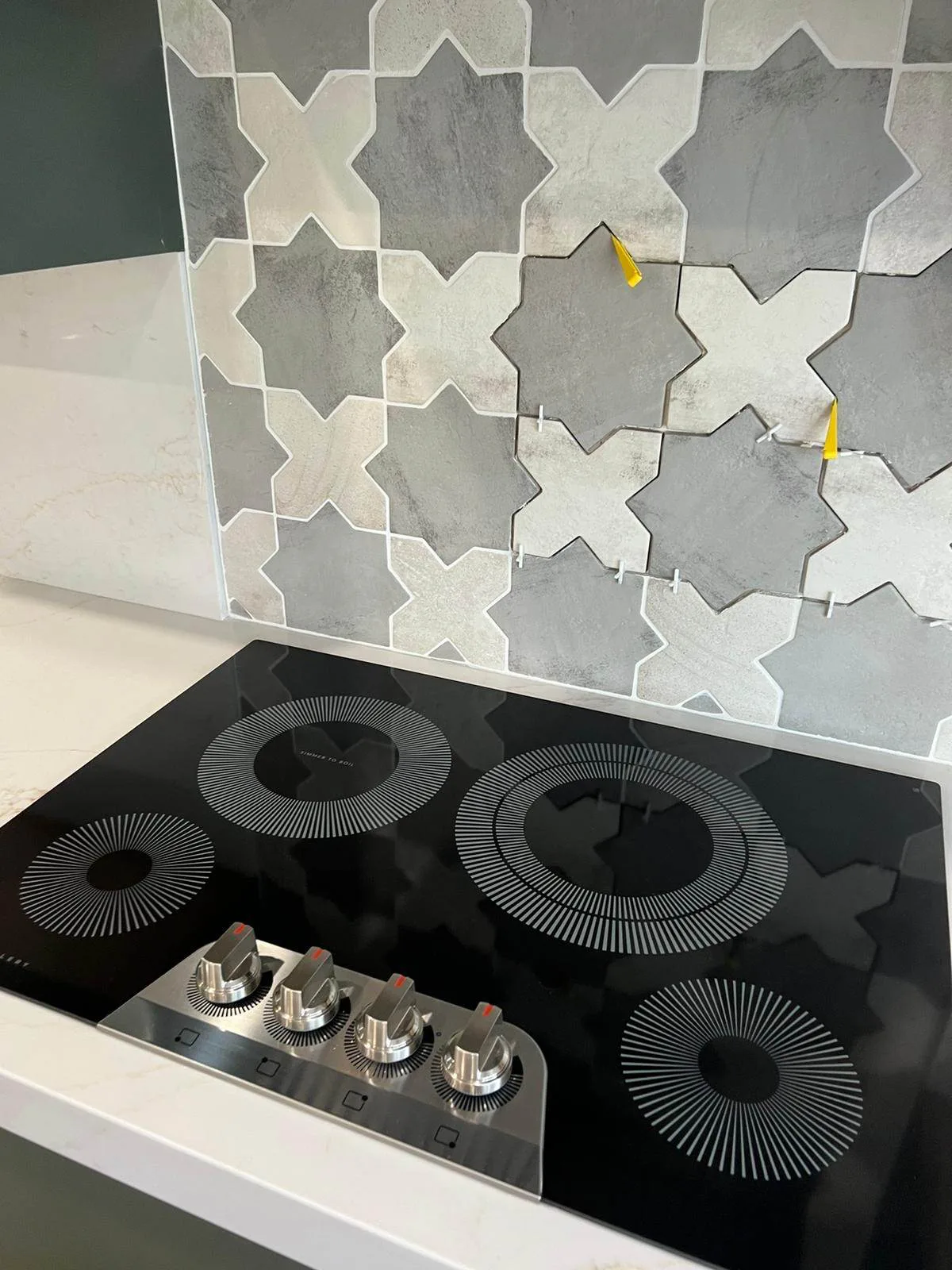 Star-and-cross pattern tile backsplash being installed above a black induction cooktop, with spacers still in place.