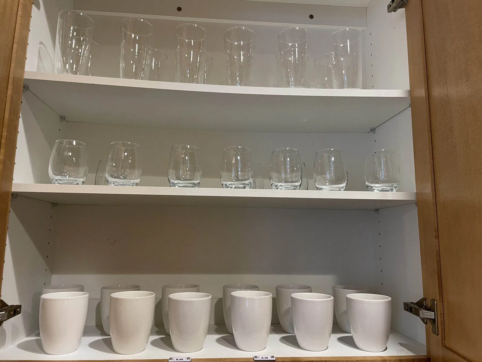 Open kitchen cabinet shelves with rows of clean drinking glasses, stemless wine glasses, and ceramic mugs.
