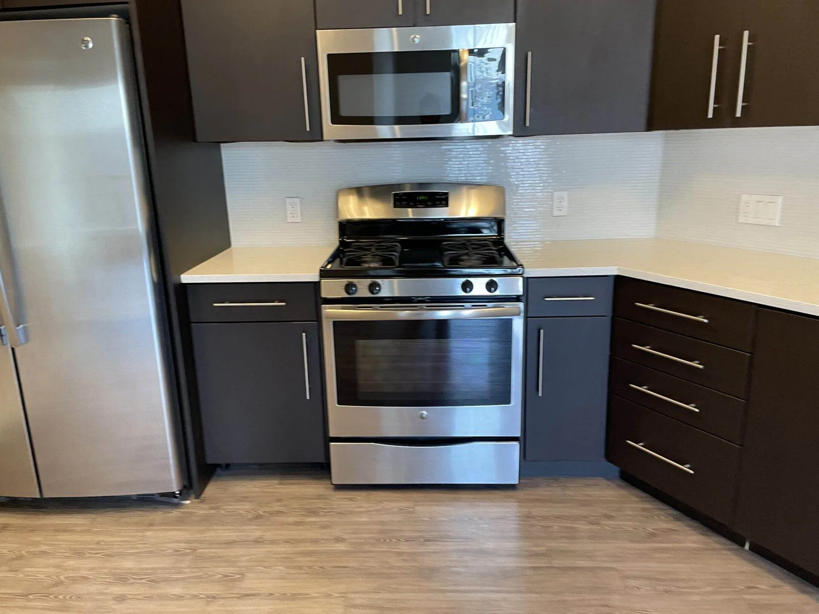 Kitchen corner with stainless steel refrigerator, gas range, and over-the-range microwave between dark cabinets.