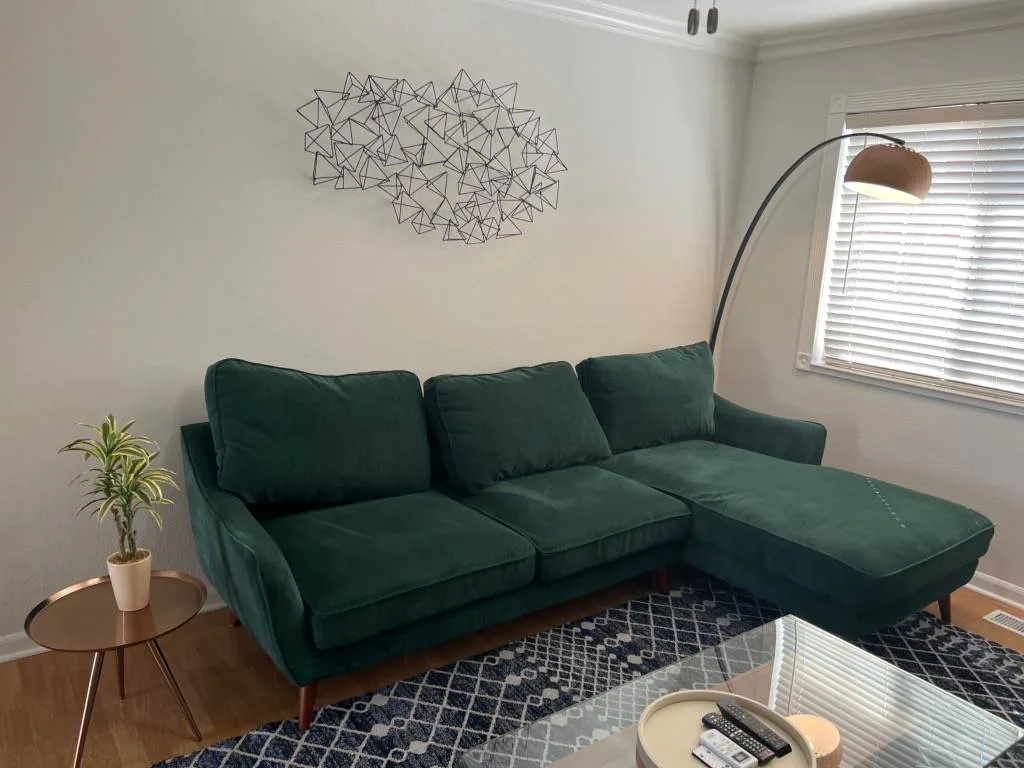 Green sectional sofa in a living room with geometric wire wall art above it, a copper side table with a small plant, and an arc floor lamp near a sliding glass door.