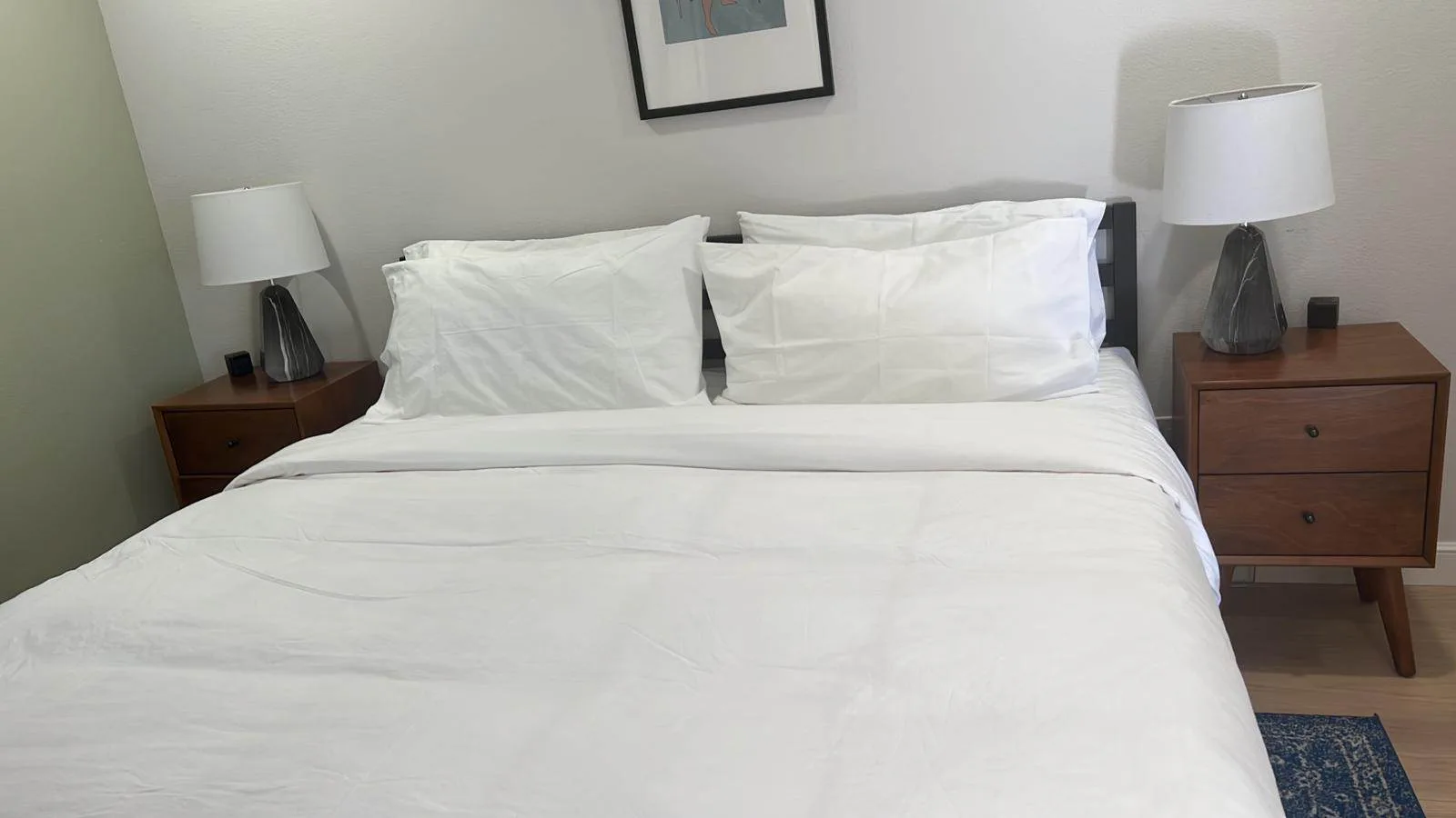 Neatly made bed with white sheets and pillows between two matching wood nightstands with lamps.