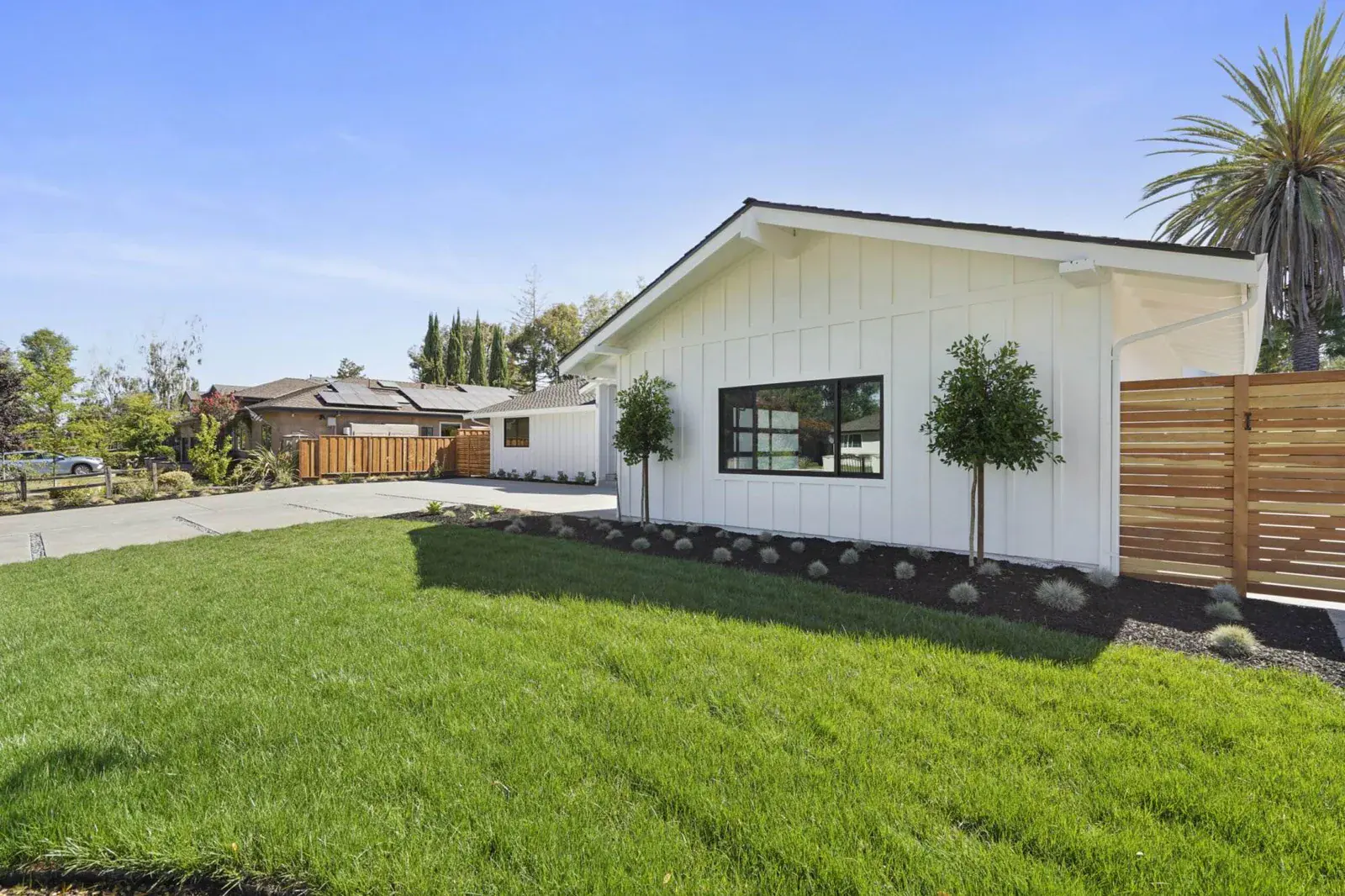 White modern farmhouse-style home exterior with black-framed window, young trees, mulched planting beds, and a wood fence.