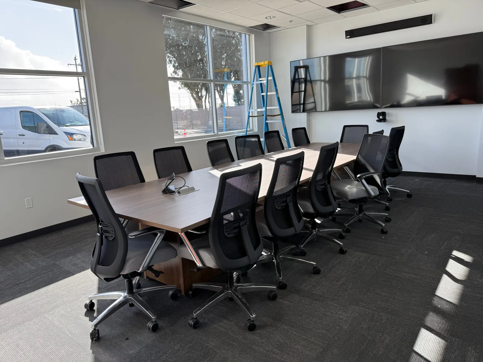 Empty conference room with a long wood table, black mesh office chairs, mounted TV screens, and a ladder by the windows.