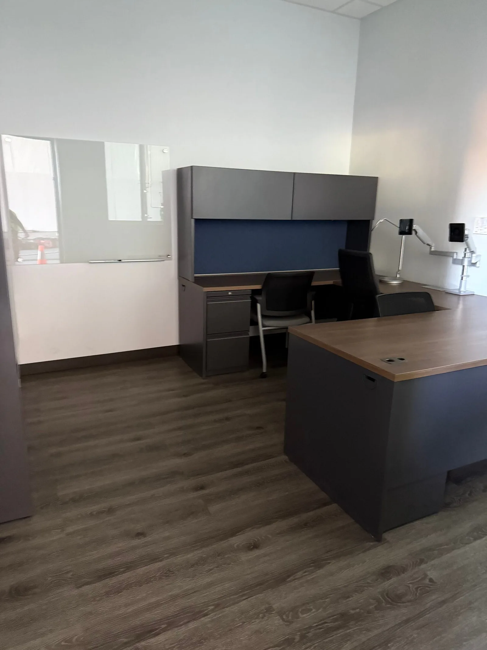 Private office with a wood-top corner desk, gray overhead file hutch, and a glass whiteboard on the wall.