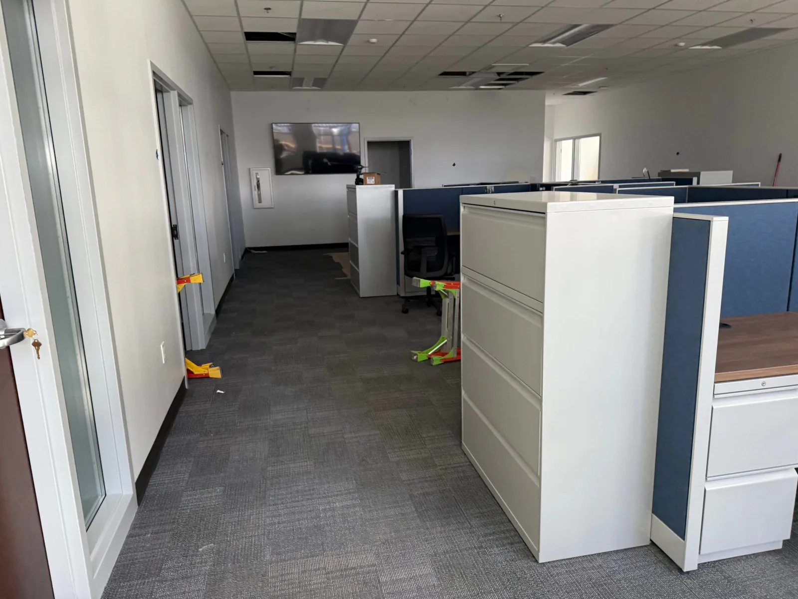 Office hallway lined with filing cabinets and cubicles, with scattered packing debris and cardboard scraps on the carpet floor.
