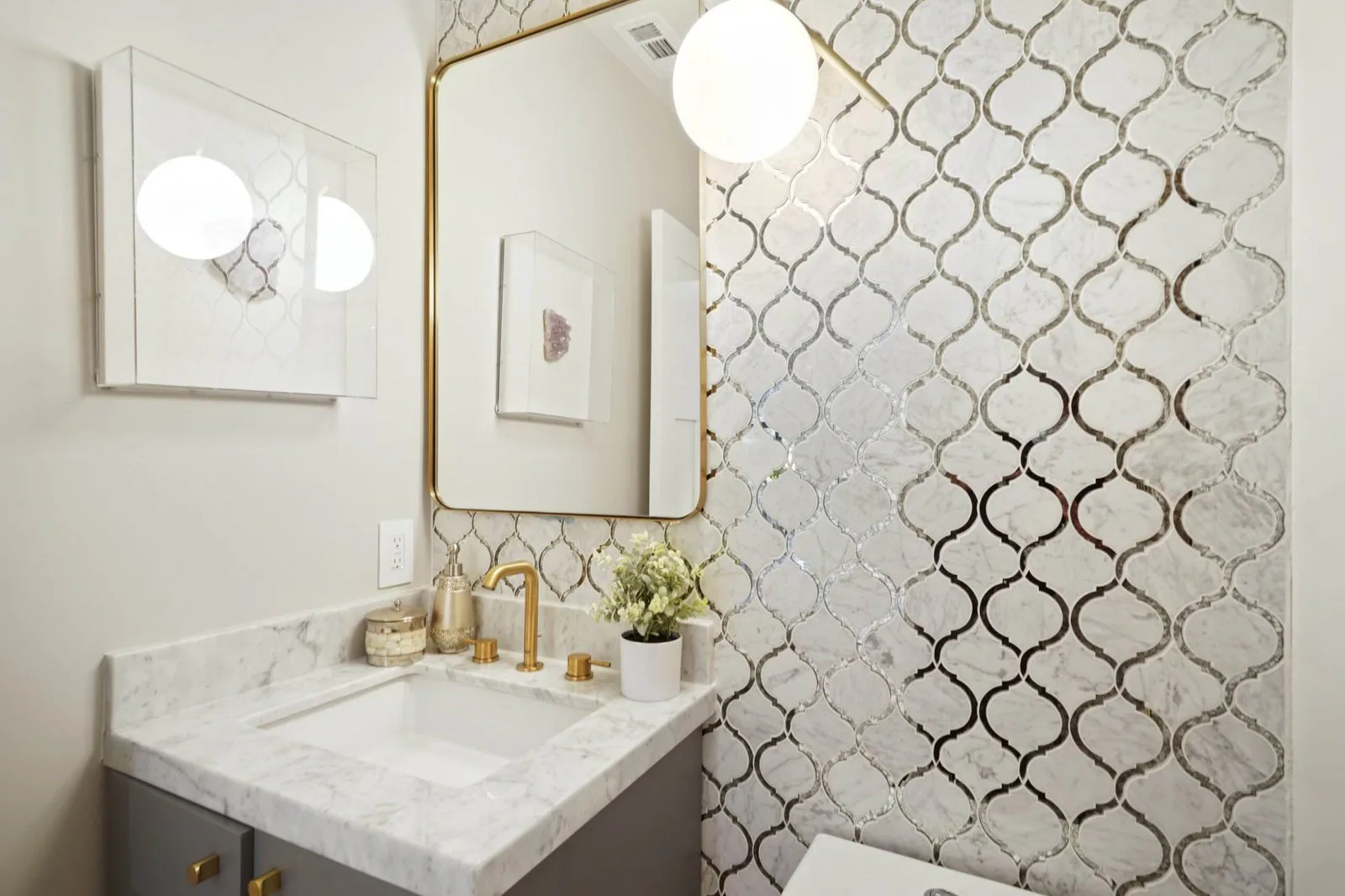 Small bathroom with silver arabesque mosaic tile, a brass-framed mirror, and marble vanity with brass faucet.