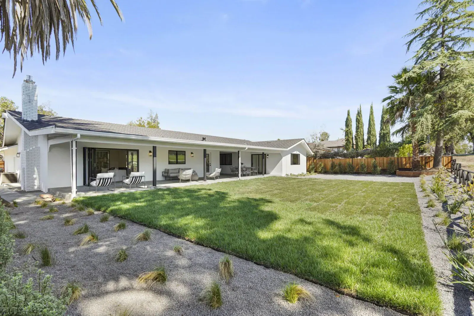 White ranch-style home exterior with a covered patio, outdoor furniture, green lawn, and tall trees against a blue sky.