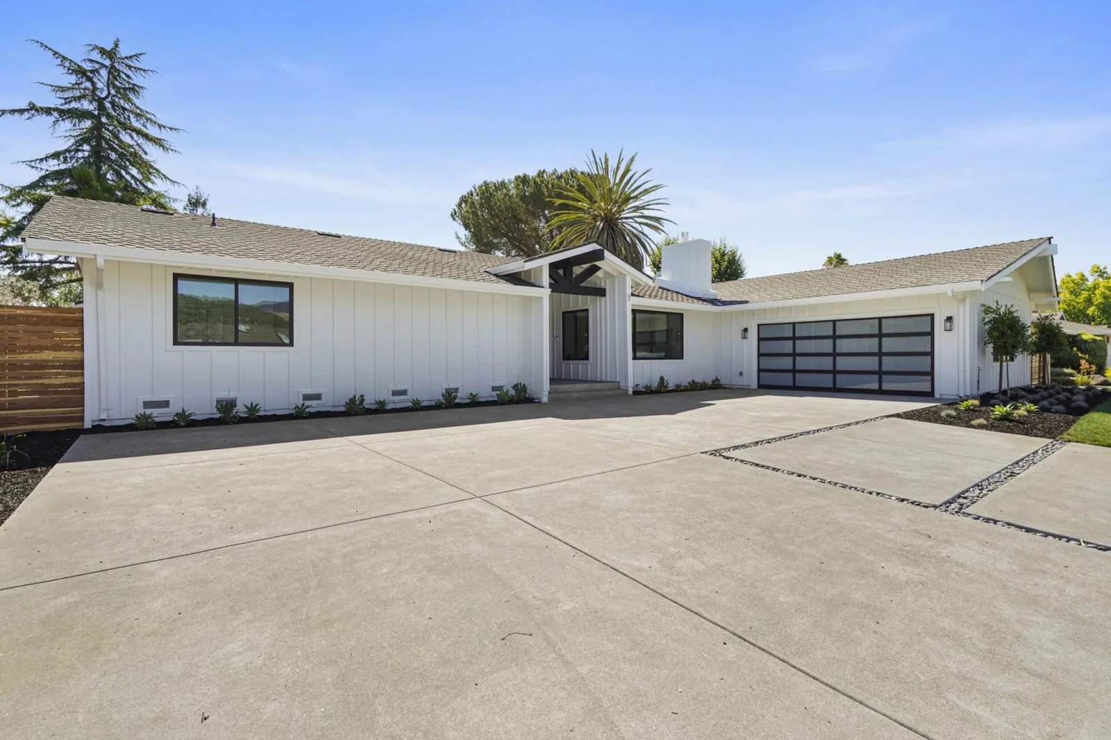 White ranch-style home exterior with a black glass garage door, concrete driveway, and palm trees against a blue sky.