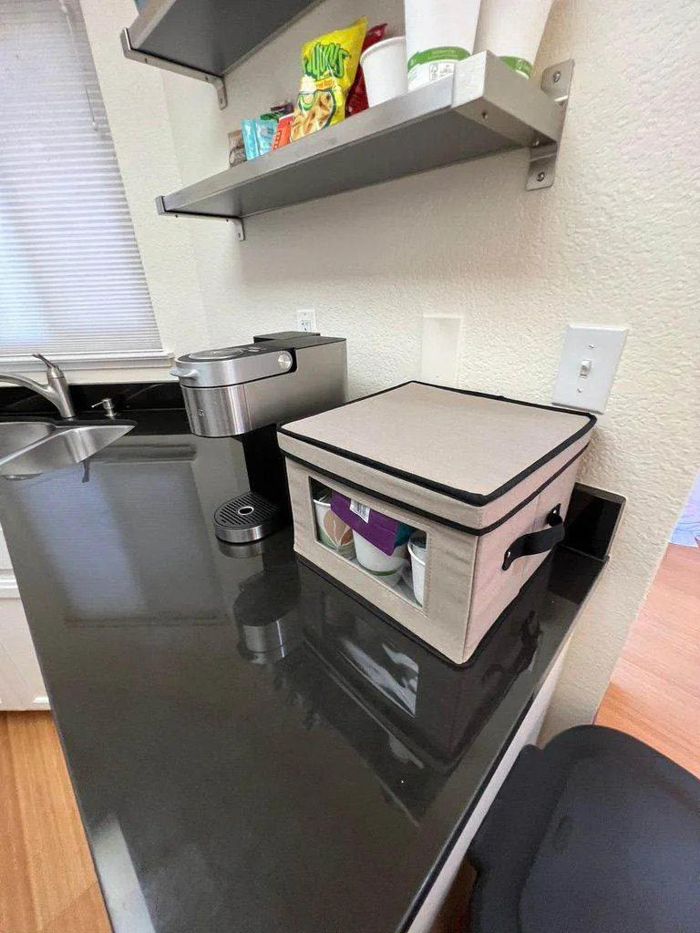Kitchenette counter with a single-serve coffee maker, snack storage bin, and stainless sink under a wall shelf.