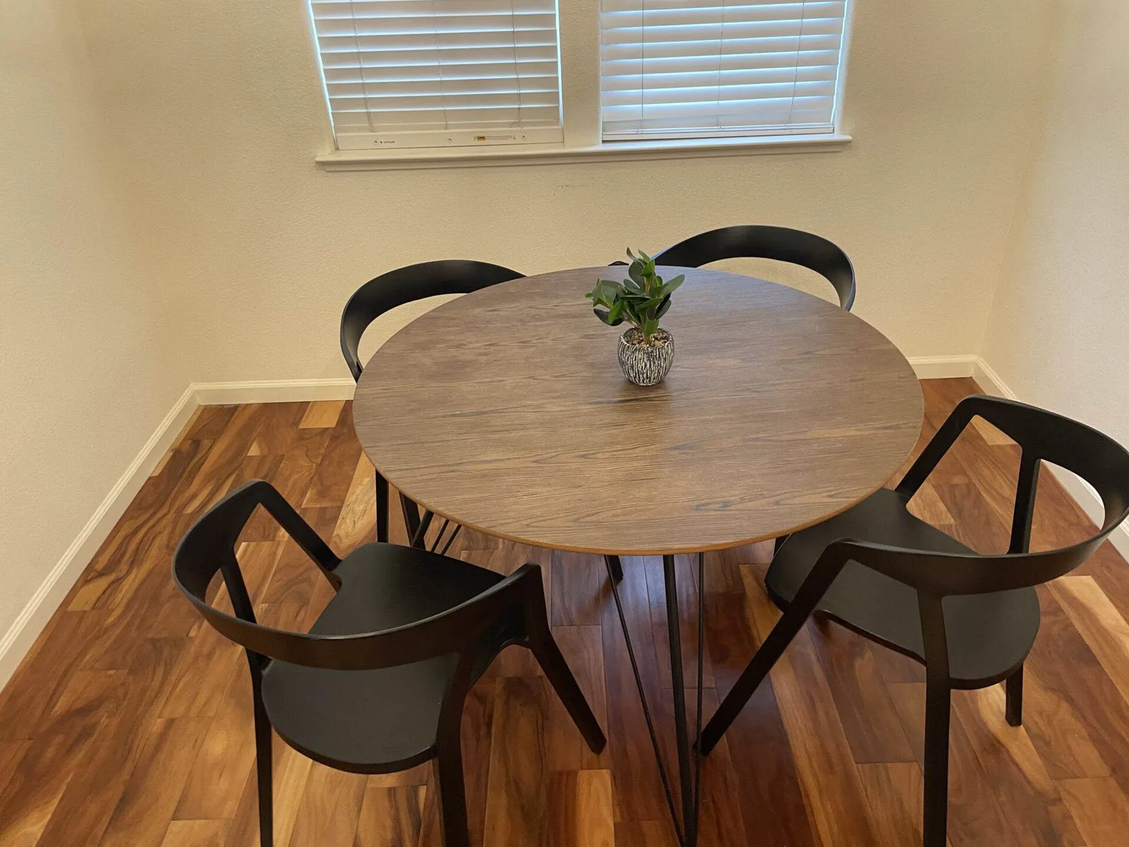 Round wood dining table with four black chairs and a small potted succulent, set in front of a window.