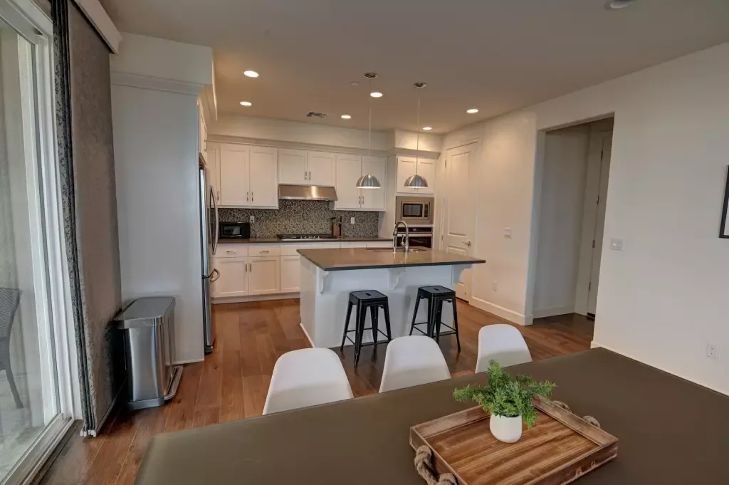 Dining table with a potted plant in front of an open white kitchen with an island and bar stools.