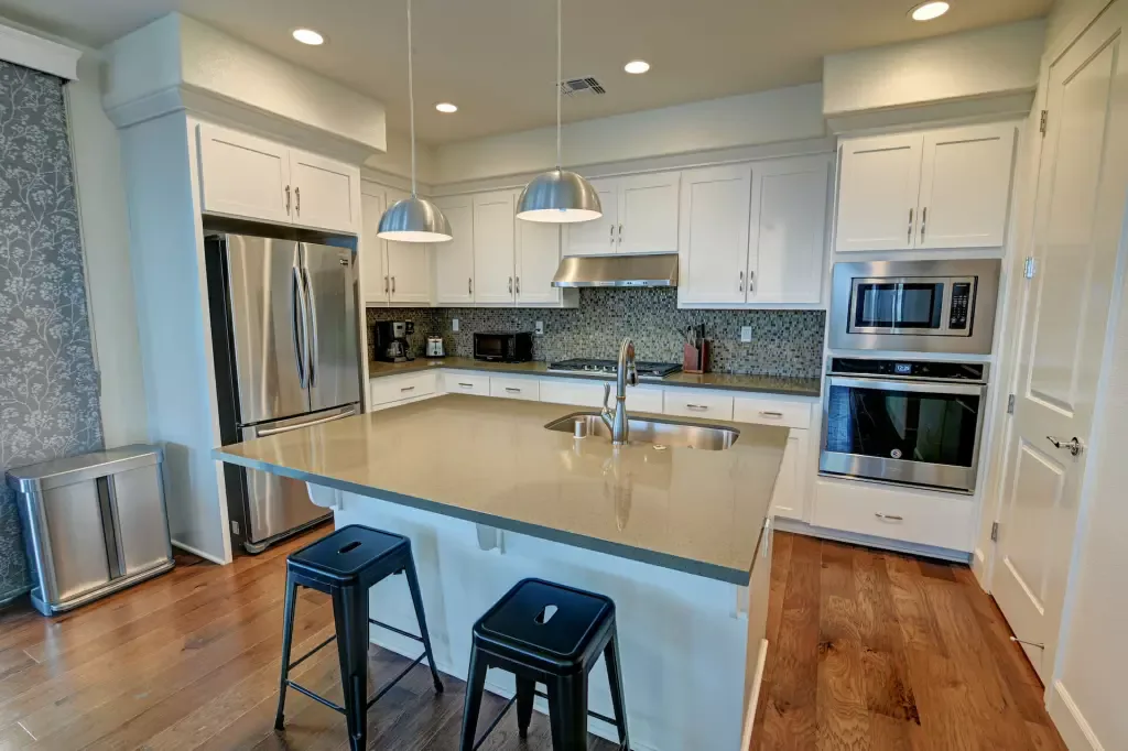 White kitchen with a large island, stainless refrigerator, and two brushed-metal pendant lights over the sink.