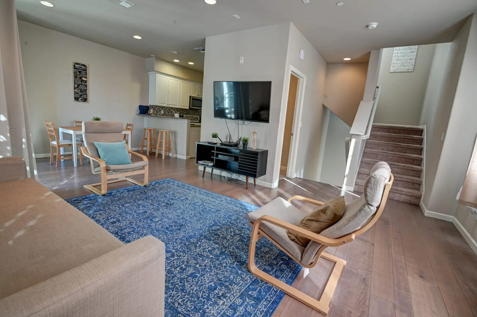 Open living room with a blue patterned rug, wood-frame accent chairs, a tan sofa, and a staircase in the background.