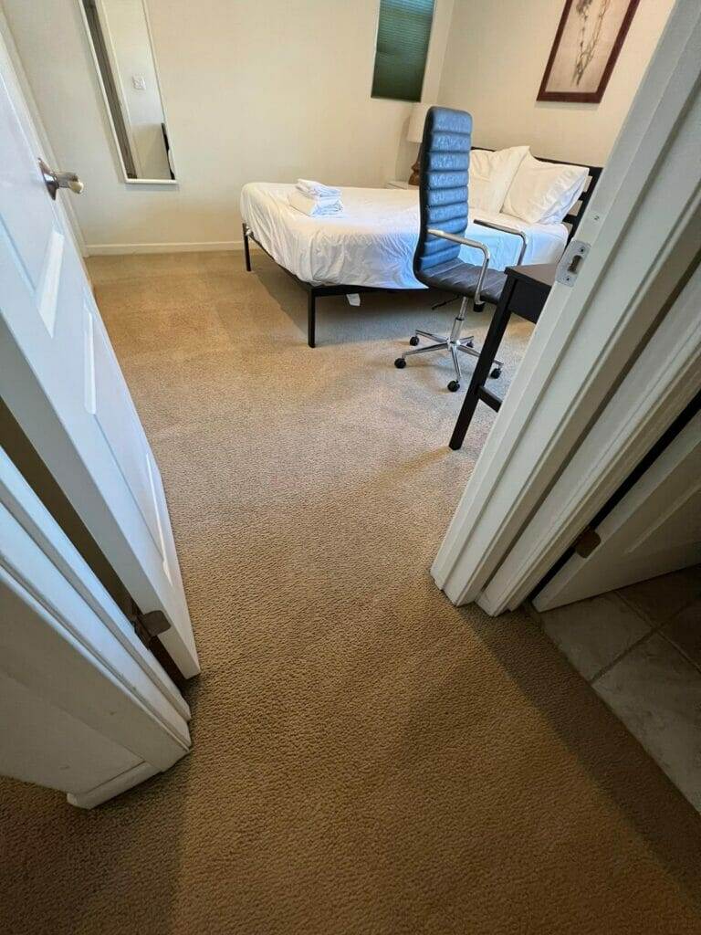 A bedroom with a bed, desk and chair in need of carpet stain removal.