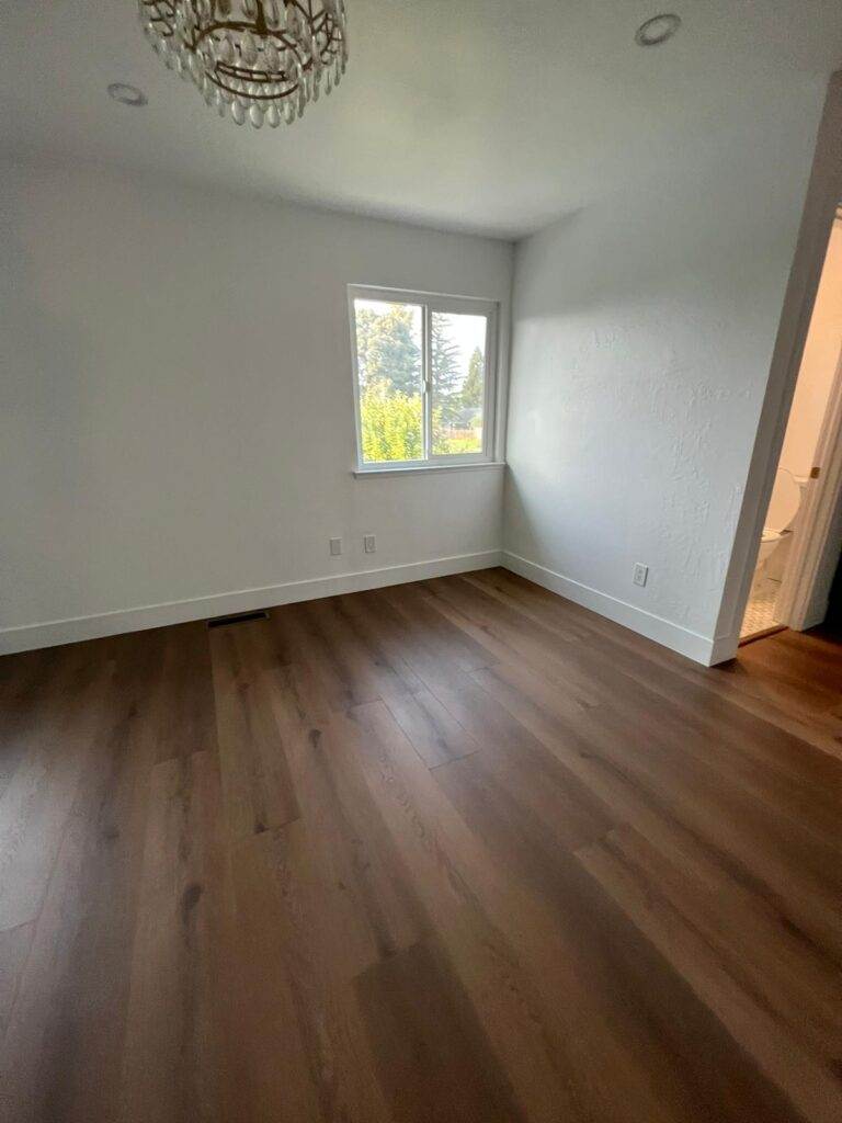 An empty room with wooden flooring, white walls, and a window providing natural light.