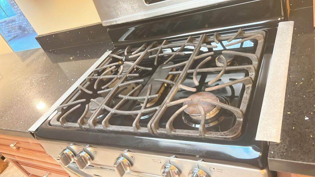 A stainless steel gas stove with four burners, one of which has visible rust, set on a granite countertop in a kitchen, ready for the professional help of Campbell Move in / Out Cleaning.