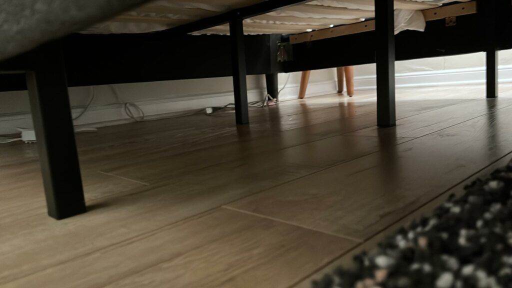 View from under a bed showing the hardwood floor, cleaned by Master Clean Service, and partial view of a person standing beyond the bed frame.