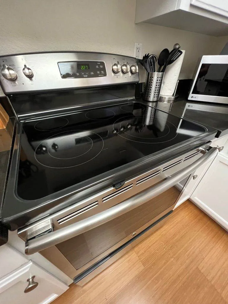 A modern kitchen featuring a black electric stove with a reflective glass top and an open oven door, surrounded by wooden cabinetry maintained by Five-Star Cleaning Experience.