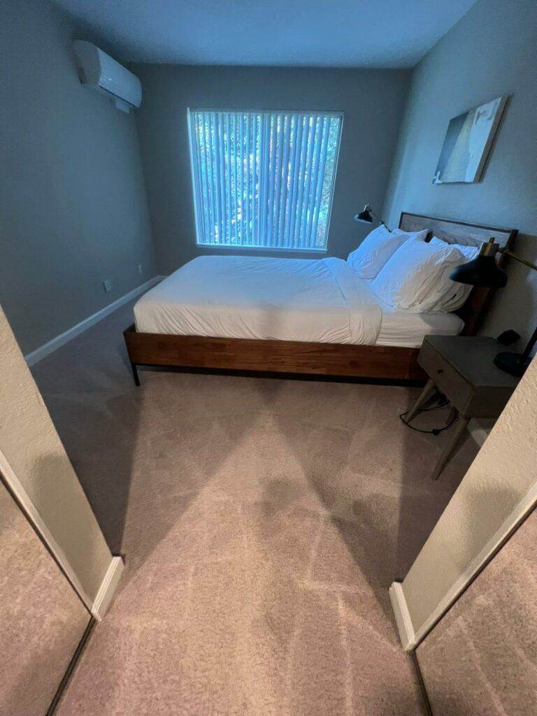 A modest bedroom featuring a large bed with white bedding, a desk, and a window with blinds, all maintained by hassle-free cleaning services and viewed from the doorway.