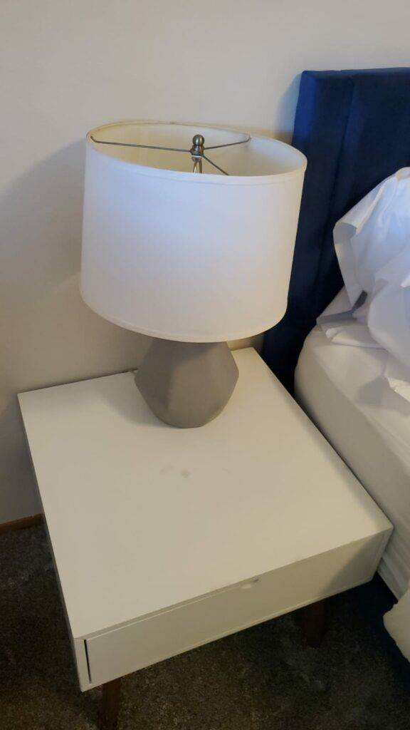 A white lamp with a cylindrical shade on a gray base, placed on a white nightstand next to a blue sofa and Master Clean Service-white bedding.