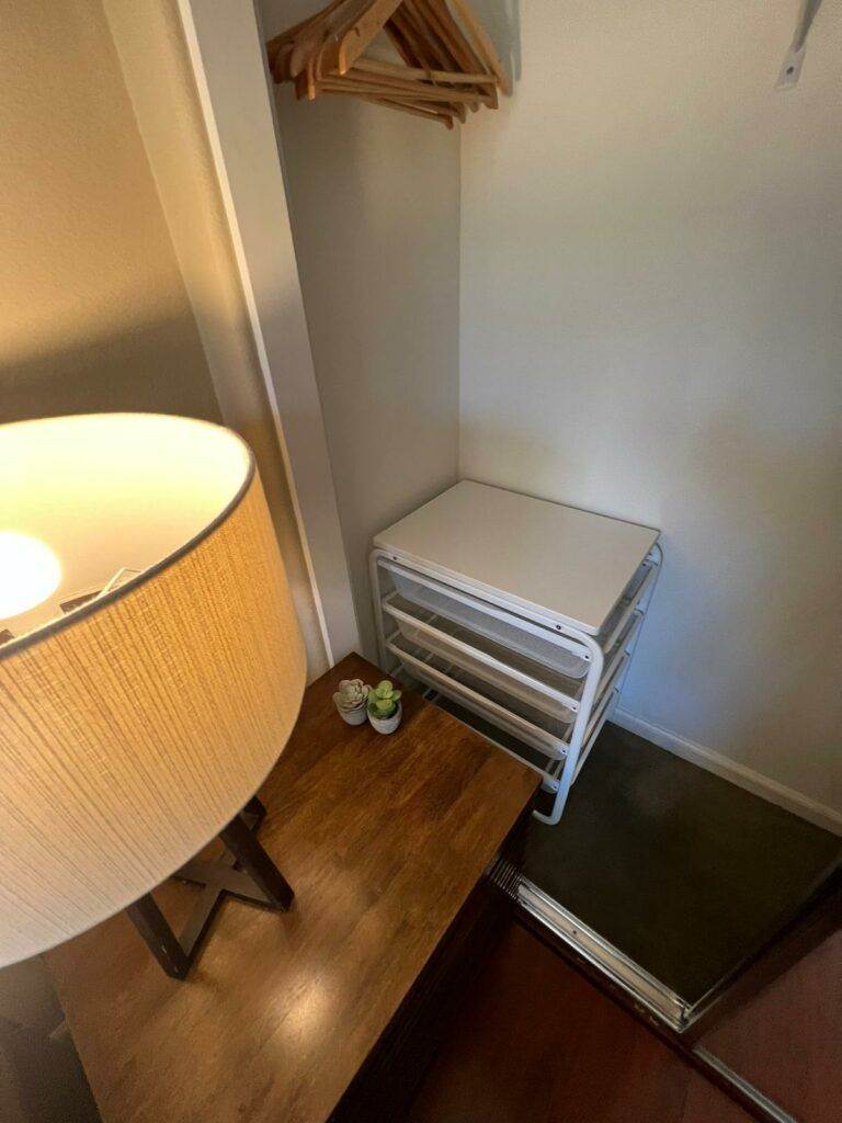 A small master bedroom corner with a wooden nightstand, a lit lamp on top, a folding luggage rack, and two small plants on the floor.