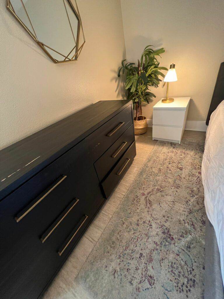 A modern bedroom with a dark dresser, white nightstand, lamp, geometric wall mirror, and a plant, complemented by a multicolored rug and maintained by Master Clean Service.