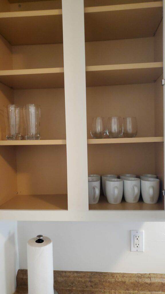 Wooden kitchen cabinet partially filled with drinking glasses, mugs, and a roll of paper towels on the countertop, maintained by Hassle-Free Cleaning.