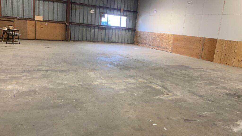 Interior of a spacious, empty warehouse with concrete flooring and wooden walls, featuring a small window and a table at the side, ready for an effortless transition.