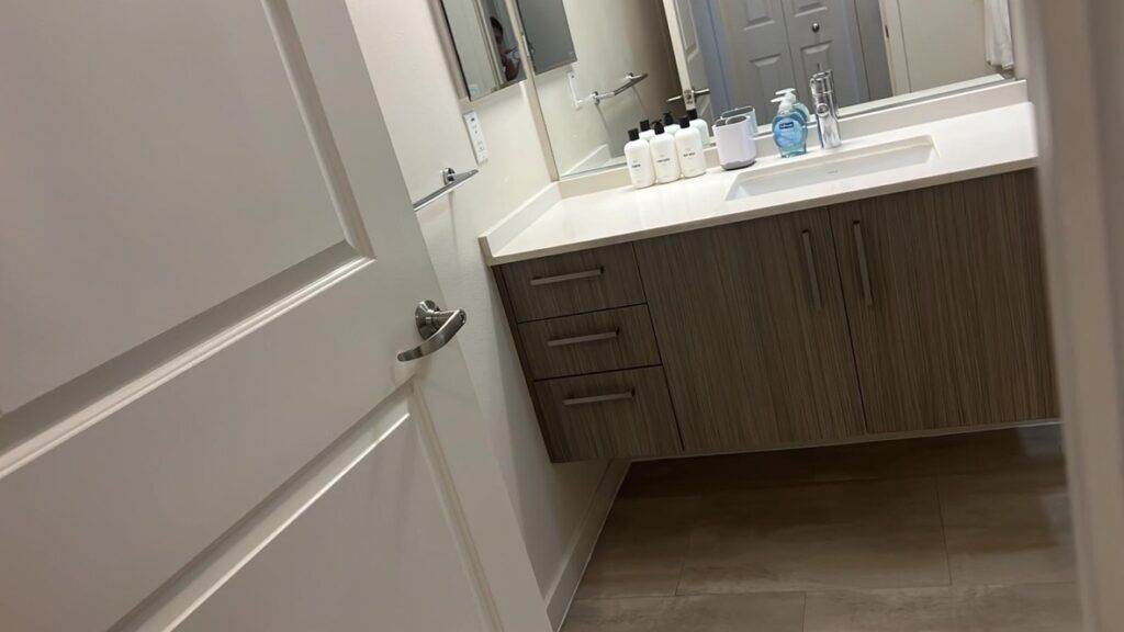 Modern bathroom with a white countertop, wooden cabinet, and two sinks with dispensers. Light tile flooring is visible, maintained by Master Clean Service.