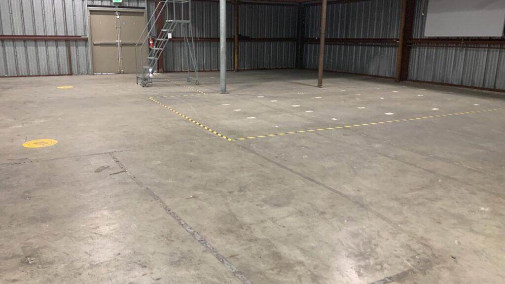 Empty industrial warehouse interior with a marked yellow boundary on the concrete floor, showing signs of a recent professional transition cleaning by Campbell Move in / Out Cleaning, and a metal ladder to one side.