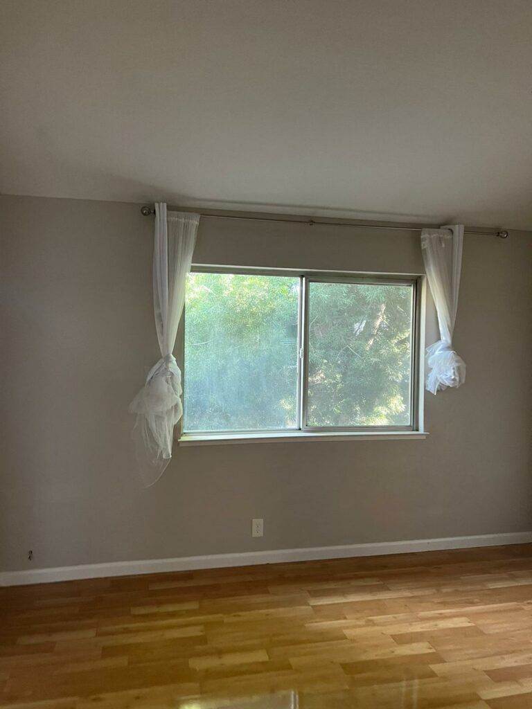 An empty room with glossy wooden floors and a large window covered with sheer white curtains, showing lush greenery outside, freshly cleaned by Master Clean Service.