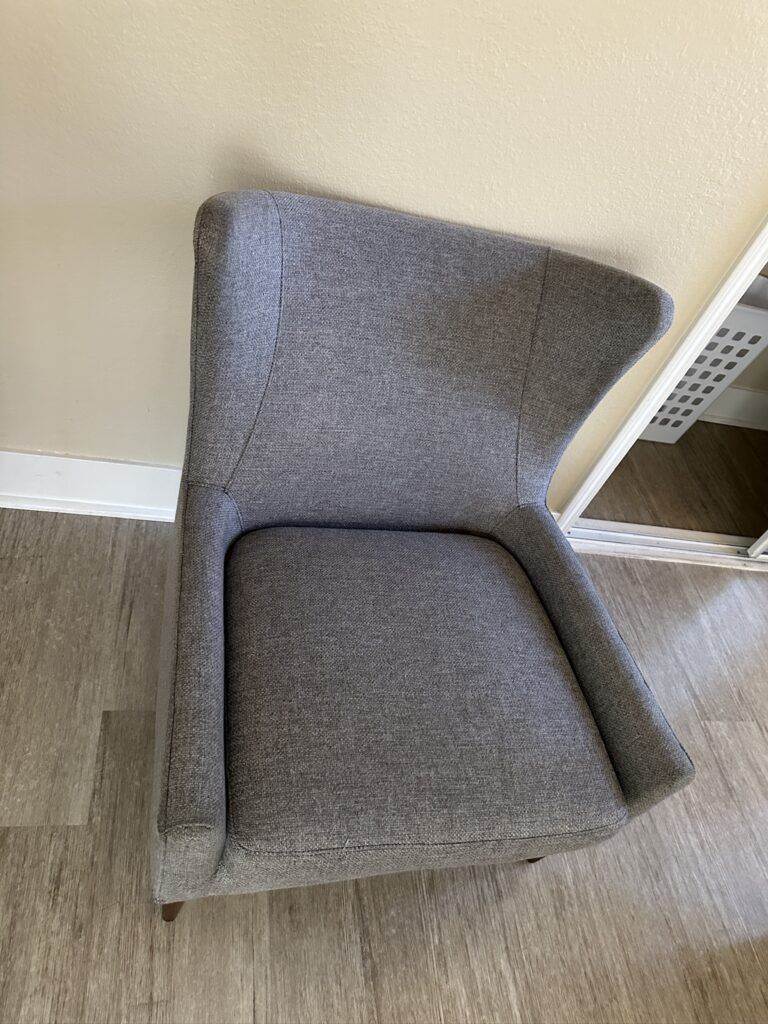 A modern grey fabric armchair with Professional Upholstery Care, featuring a high back and angled arms, standing on a light wooden floor against a cream wall.