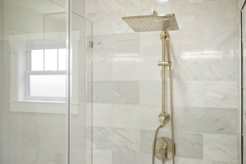 Modern shower with a gold-colored square shower head, mounted in a white marble tiled enclosure from our construction portfolio, featuring a small window.