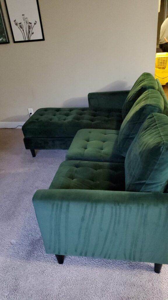 A green velvet sectional sofa, one of the upholstered pieces in a living room with light gray carpet, and framed wall art visible in the background.