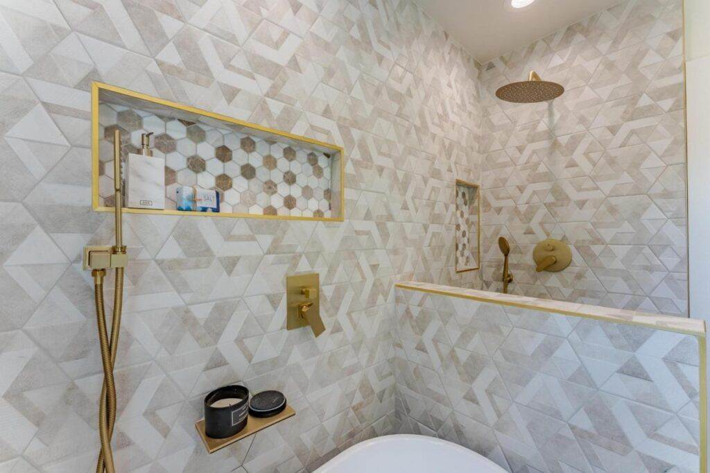 Modern bathroom with geometric tile patterns, gold fixtures, a wall-mounted shelf with toiletries, and a large showerhead, perfect for a post-construction portfolio.