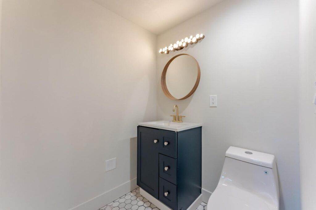 A small bathroom featuring a dark grey vanity with drawers, a round mirror above it, and a white toilet to the right, illuminated by a row of lights above the mirror, recently added to our post