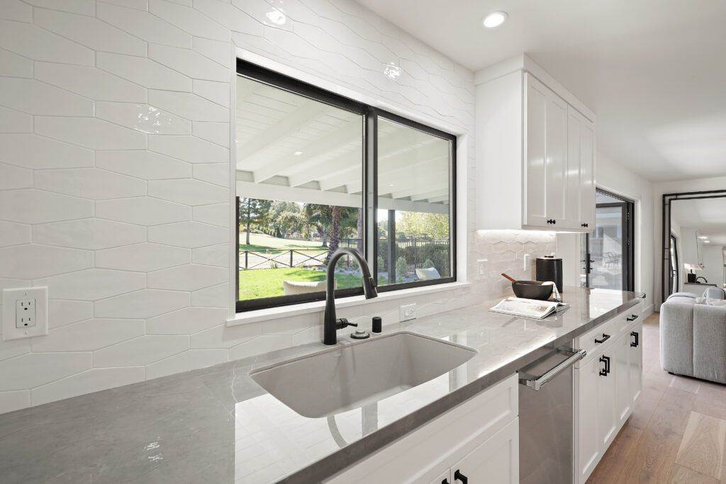 Modern kitchen with white subway tiles, stainless steel sink, large windows overlooking a green yard, and white cabinetry, newly completed in our construction portfolio.