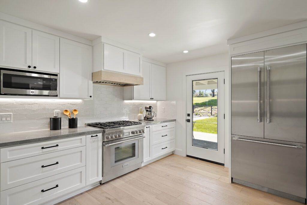 Modern kitchen interior with white cabinetry, stainless steel appliances, and wooden flooring showcased in our construction portfolio, with natural light coming in through an open door.