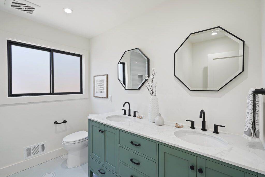 Modern bathroom with teal cabinets, dual sinks, octagonal mirrors, and a large window showcasing a sunset view, featured in our post-construction portfolio.